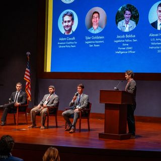 Three panelists seated on stage and a speaker at the podium, beneath a slide with the speakers' photos and titles