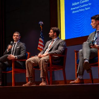 Three panelists on stage, the panelist on the left speaking into a microphone while the other two listen